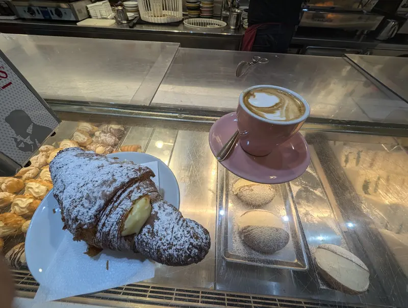 Cornetto alla crema con zucchero a velo e cappuccino in tazza rosa sul banco del bar